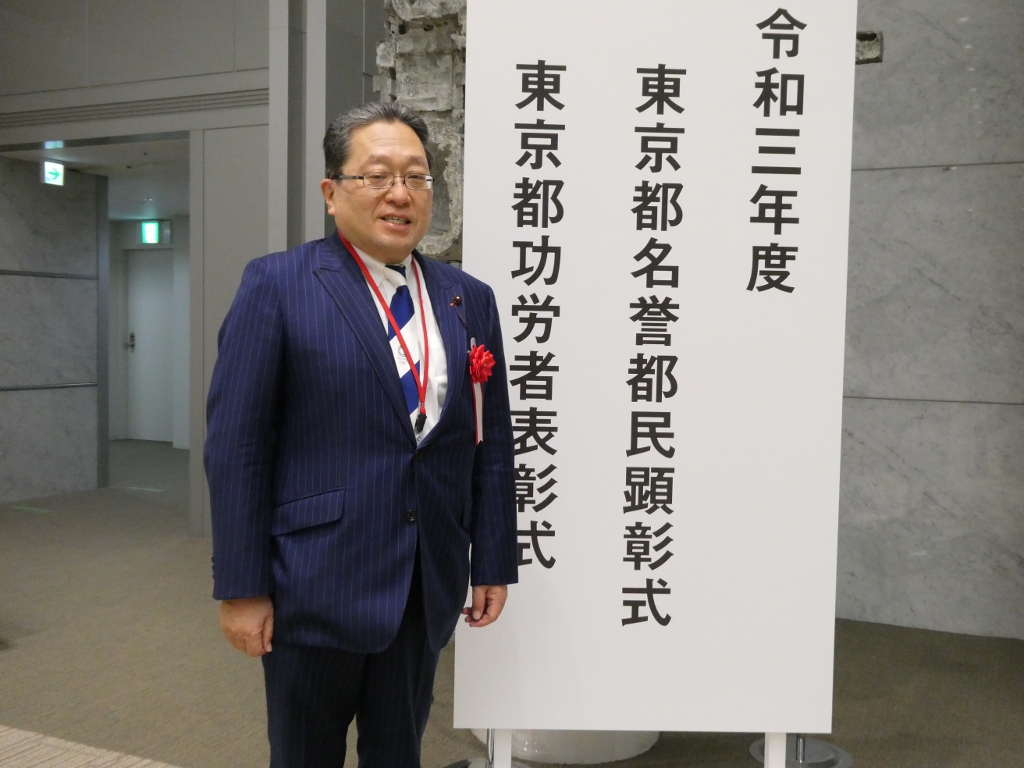 写真：東京都庁舎の表彰式会場看板横に立つ丸山議員の様子