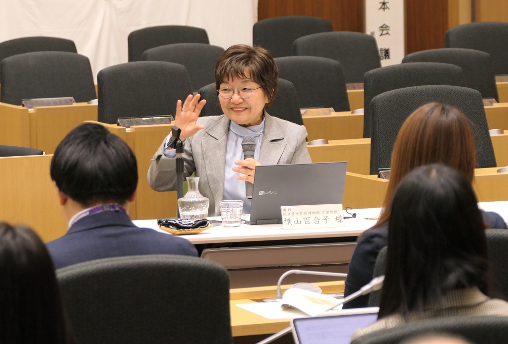 写真：議場で席に着いて話す　講師の　よこやま ゆりこ氏